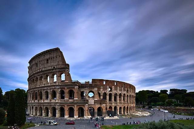 The Colosseum at dawn under a streaked blue and violet sky, with the road and the square in front